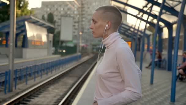 Mujer caucásica sin pelo está mirando a la estación de tren y se mueve en la toma de la música en los auriculares a la luz del sol — Vídeos de Stock
