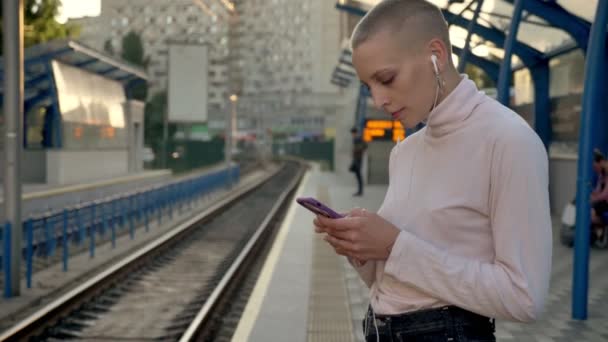 Agradable calvo confiado chica caucásica está mirando a los trenes en la estación de metro, vista urbana, fondo soleado — Vídeo de stock