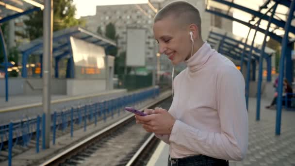 Hermosa mujer calva joven está de pie y se mueve en la danza en la estación de tren, deslizando el teléfono con sonrisa, fondo de la ciudad — Vídeos de Stock