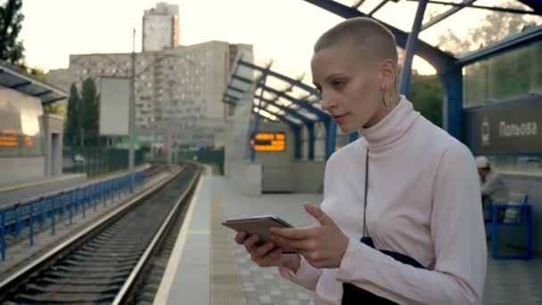 Junges haarloses Mädchen beim Blick auf ihr Tablet und bei der Kontrolle von Zügen am Bahnsteig tagsüber in der Stadt — Stockvideo