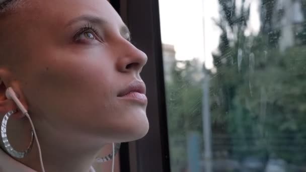 Mooie close-up gezicht blond Kaukasische verdrietig meisje glazen venster in trein kijken en luisteren naar muziek alleen — Stockvideo
