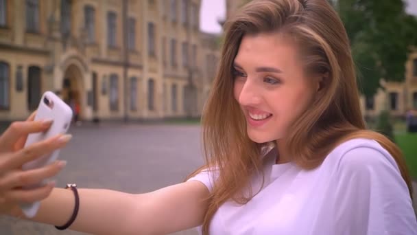 Tímida y bonita chica caucásica con el pelo largo tomando selfies con su dispositivo en la vista urbana, día soleado — Vídeo de stock