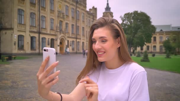 Cute caucasian confident female is having phone conversation with camera, talking emotionally and smiling with street and buildings behind her — Stock Video