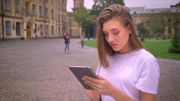 Офигенная блондинка-кавказка печатает на планшете с концентрированным лицом, стоя на улице со зданиями на заднем плане — стоковое видео