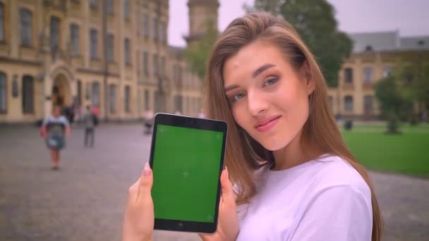 Jolie fille caucasienne montre écran vert et souriant heureux et détendu sur la vue urbaine, de jour — Video