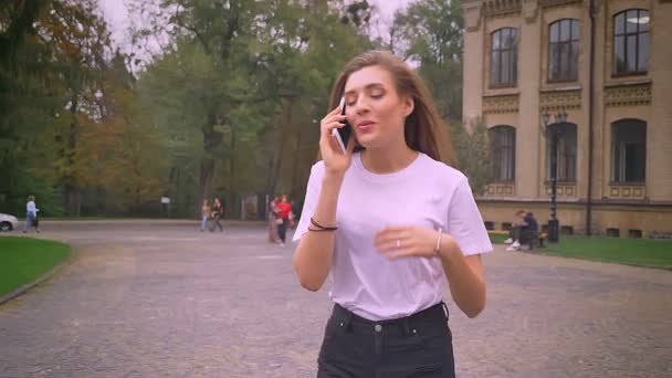 Bonito caucasiano menina está falando por telefone com sorriso enquanto caminhando no parque sozinho, durante o dia, movimento rápido — Vídeo de Stock