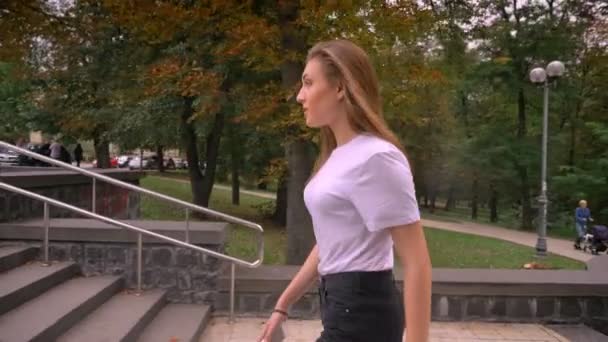 Bella ragazza caucasica sta camminando nel parco, attraversando la strada con i capelli lunghi che volano nel vento — Video Stock