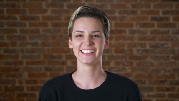 Close-up portrait of laughing cute caucasian short-haired blonde girl, standing and looking at camera on brick wall — 图库视频影像