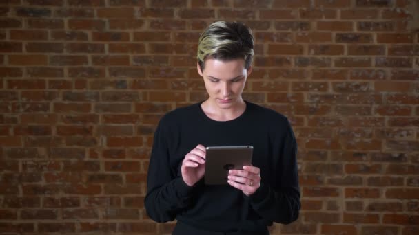 Mulher muito caucasiana com cabelo curto está passando seu tablet enquanto está isolada ao lado da parede de tijolo em suéter preto — Vídeo de Stock