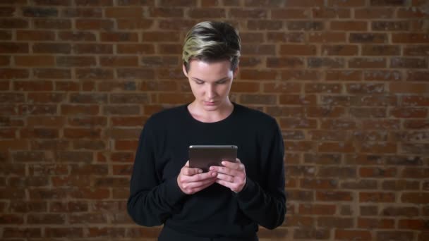 Mulher branca bonita está usando seu tablet e sorrindo encantador para a câmera no fundo de tijolo isolado — Vídeo de Stock