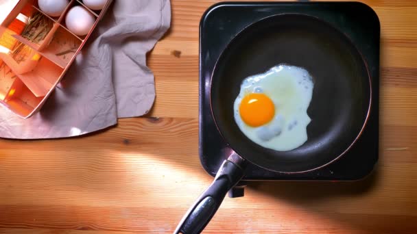Выстрел сверху вниз, одно жареное яйцо на сковороде становится золотым и готовится к еде, деревянный стол в солнечных лучах — стоковое видео