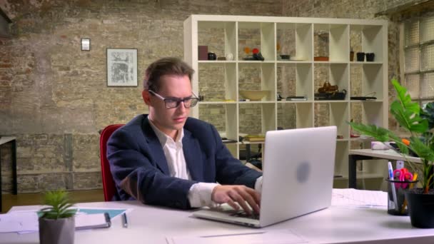 Caucasiano satisfeito em sua mesa de trabalho usando laptop enquanto sentado com tijolo verde atrás — Vídeo de Stock