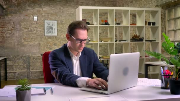 Gelukkig Kaukasische zakenman winnende teken met handen en houd waarop werken bij zijn laptop in lichte baksteen kantoor geïsoleerd — Stockvideo