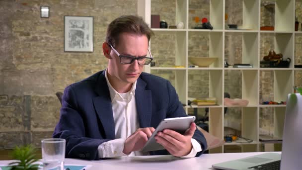 Focused caucasian businessman is scrolling his tablet and drinking water while working at his desk inside brick studio — Stock Video