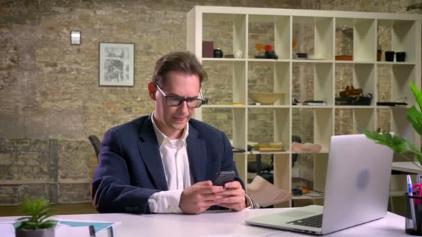 Глибоко зосереджений білий бізнесмен прокручує свій смартфон, сидячи за білим столом в цегляному офісі — стокове відео