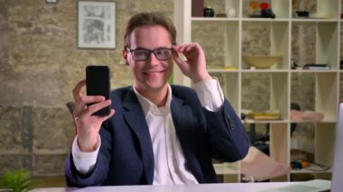 Sevimli beyaz erkek takım elbise, onun smartphone, office ruh selfie çekerken gözlüğünü tamir ediyor