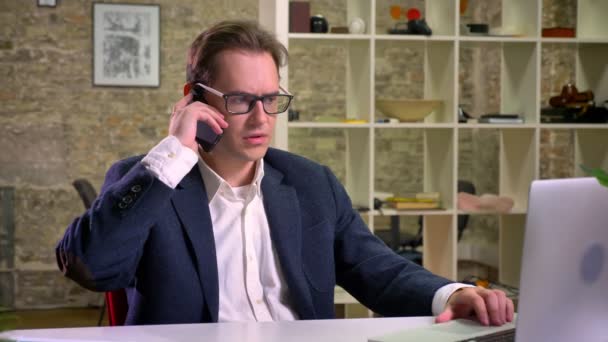 Serious caucasian man is having phone conversation while sitting near laptop in brick office isolated, classic suit and glasses — Stock Video