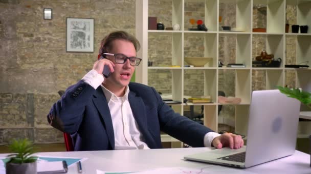 Energetic talking caucasian man in business suit and glasses hanging on the phone and talking relaxed while sitting at white working desk indoor — Stock Video