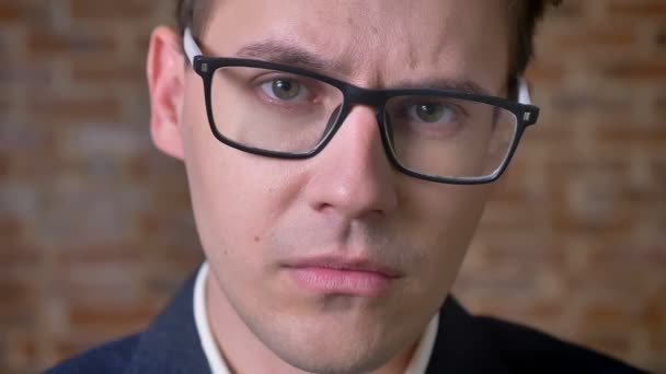 Close-up cara entediada de homem de negócios caucasiano em óculos pretos olhando em linha reta e confiante no estúdio de tijolos — Vídeo de Stock