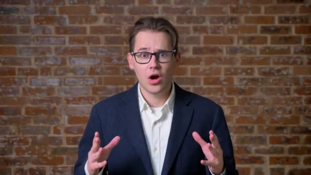 Enorme sorpresa en la cara de hombre de negocios caucásico que está de pie y la fijación de sus gafas en el estudio de ladrillo — Vídeo de stock