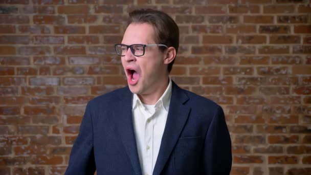 Yawning caucasian guy is standing next to camera on brick background and looking tired and relaxed indoor — Stock Video