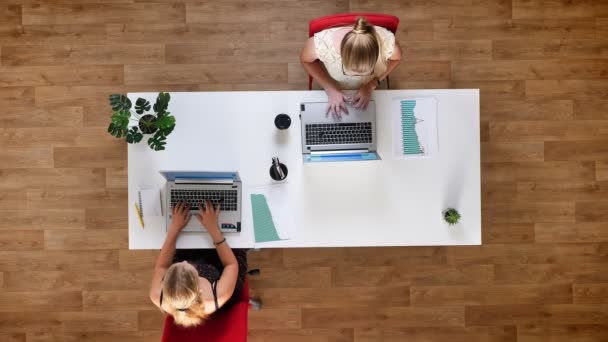 Сверху вниз выстрелили продуктивные коллеги, две девушки сидят за общим столом и работают на своих ноутбуках на деревянном полу, в помещении — стоковое видео