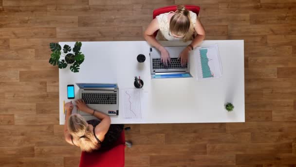 Top-down skott, två produktiva flickor sitter vid bordet i trä office och använder sina bärbara datorer och blå skärm smartphone — Stockvideo