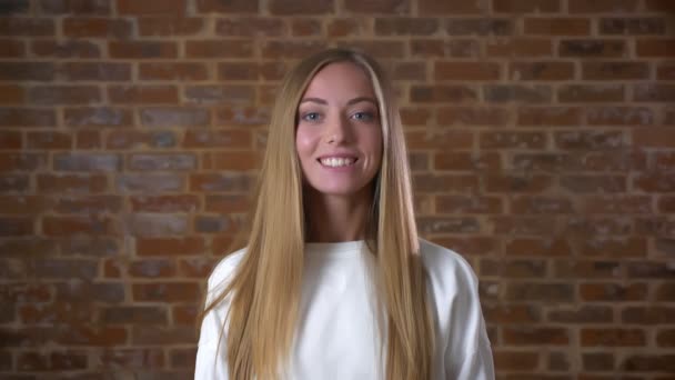 Hermosa chica caucásica sonriente en el estudio de ladrillo de pie recto y sonriendo a la cámara con encanto — Vídeos de Stock