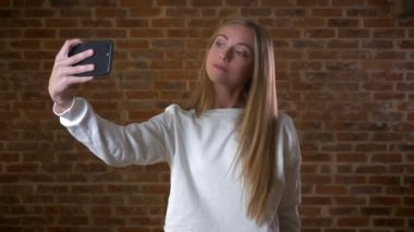 Sarışın beyaz kız selfie alıyor ve seyir onun telefon gülümseme ve mutlu yem yüz, ile izole kırmızı tuğla duvara