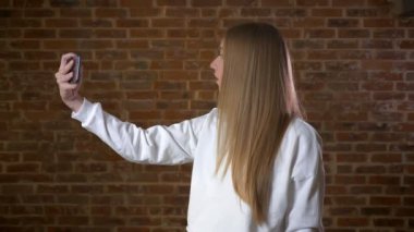 Yakışıklı beyaz sarışın kadın profil tuğla Studio tek başına rahat ayakta onu telefonunuzun web kamerada fotoğraf çekmek