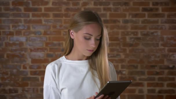 Красива біла жінка прокручує свій планшет зі сфокусованим обличчям і дивиться на екран, стоячи на червоному цегляному тлі — стокове відео