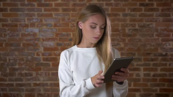 Awesome caucasian girl typing on her tablet with high concentration and standing next to camera in red studio near brick — Stock Video