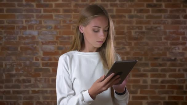 Pretty blonde caucasian girl is smiling at camera and swiping the tablet chilled and amused, indoor, red brick studio — Stock Video