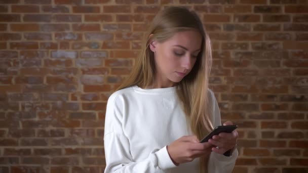 Nice blonde caucasian girl in scrolling her smartphone and looking precisely while standing next to the brick wall indoor isolated — Stock Video