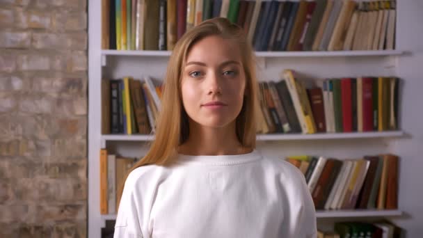 Frisches, schönes Gesicht einer kaukasischen Frau, die mit einem Lächeln in die Kamera blickt und allein in der Bibliothek steht — Stockvideo