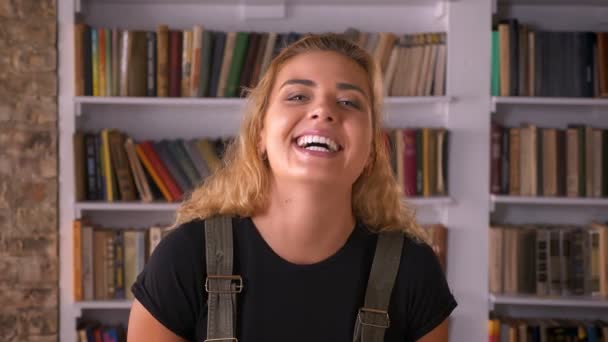 棚の本の横に立っている間カメラでかわいい誠実な白人金髪女性が笑っています。 — ストック動画