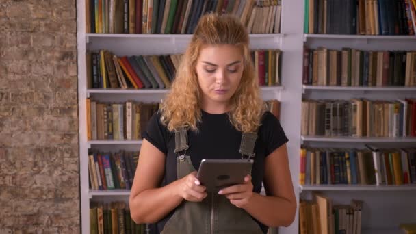 Awesome caucasian curly red-headed girl is swiping tablet and smiling at camera happily with books behind her — Stock Video