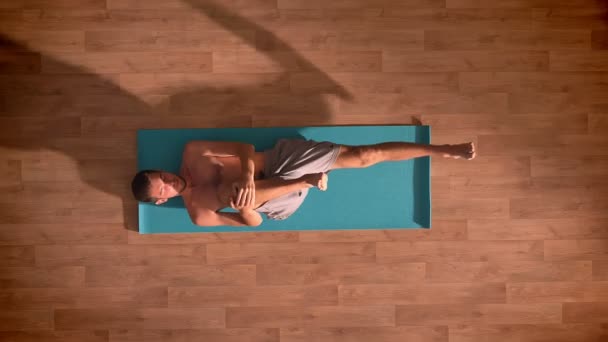 Tiro superior, homem caucasiano está deitado em um tapete de ioga e esticando as pernas com os olhos fechados no chão de madeira — Vídeo de Stock