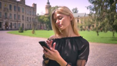 Çok beyaz sarışın kadın telefonunu swiping ve sokakta rahat dururken kullanma