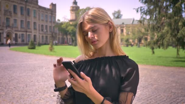 Aranyos kaukázusi szőke lány a görgetést a smartphone állva, az utca közepén, és nyugodtan mosolyogva — Stock videók