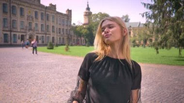 Enjoyable dancing blonde female with satisfied look hanging out on the street near park, city background