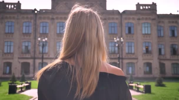 Tir par derrière de fille caucasienne blonde qui sort de la caméra, prenant la direction du bâtiment, à l'extérieur — Video