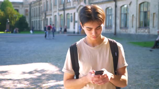 Hnsome sorrindo asiático homem está rolando seu telefone enquanto está na rua, raios de sol e edifícios antigos atrás dele — Vídeo de Stock