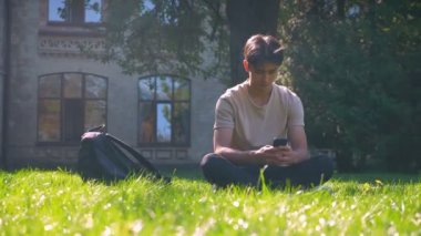 Asya yakışıklı sunlights çimenlere oturup ve vibes eğitim onun telefon, odaklanmış, kentsel görünümü kaydırma