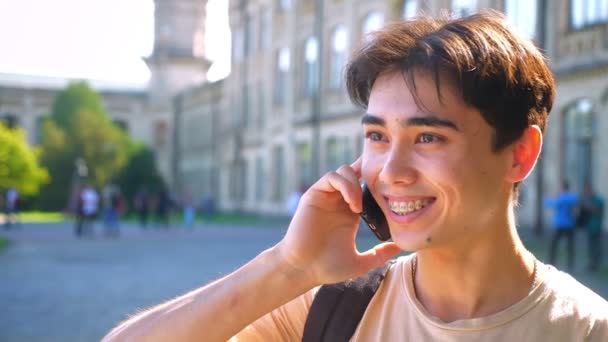 Nice asian boy is on the phone, smiling and talking, bright sun, urban view as background — Stock Video