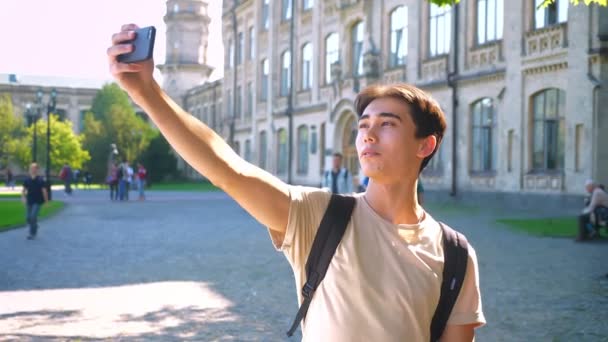 Awesome asian boy is taking selfie with his smartphone while standing near ancient building in sunlights — Stok Video