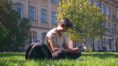 Şirin çim Asya'ya ait adam Park'ta oturuyor onun smartphone, daire ile fotoğraf makinesi, gülen yüz, sakin ruh gülümseyen shooted kullanıyor