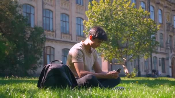 Χαριτωμένο κάθεται στο πάρκο στον ασιατικό τύπο χλόης είναι χρησιμοποιώντας το smartphone του, τεράστιο κύκλο με φωτογραφική μηχανή, χαμογελώντας χαρούμενο πρόσωπο, ψύχρα διάθεση — Αρχείο Βίντεο
