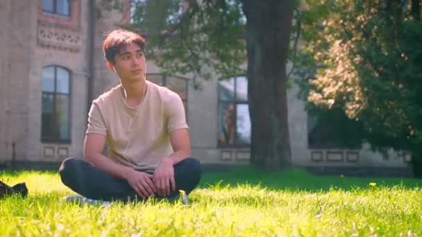 Agradable asiático chico es involucrado en escuchar música en auriculares mientras sentado en la hierba con edificios detrás, aislado — Vídeos de Stock
