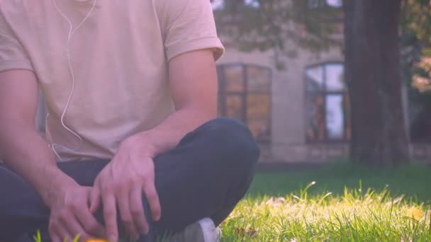 Relajado asiático hombre es escuchar música mientras colgando fuera en parque, sentado en la hierba, antiguo ciudad — Vídeo de stock